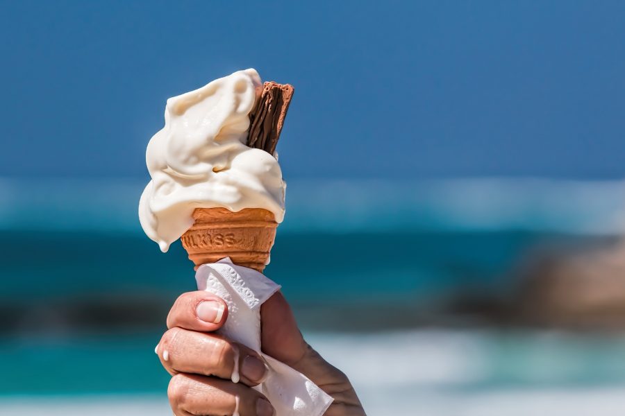 Helado de cucurucho con nata y chocolate sujetado por la mano de una mujer en la playa.